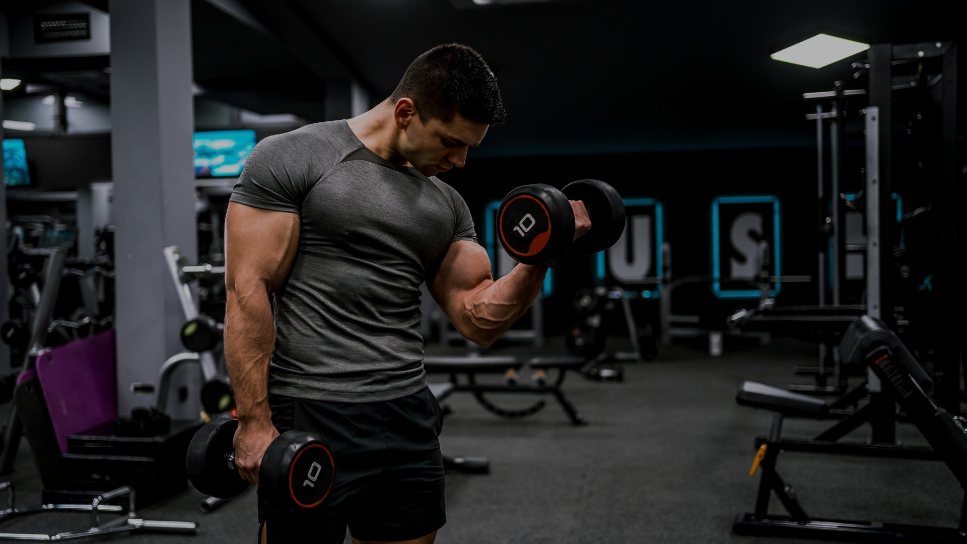 A muscular man in a gym setting, wearing a dark gray t-shirt and black shorts while lifting dumbbells.