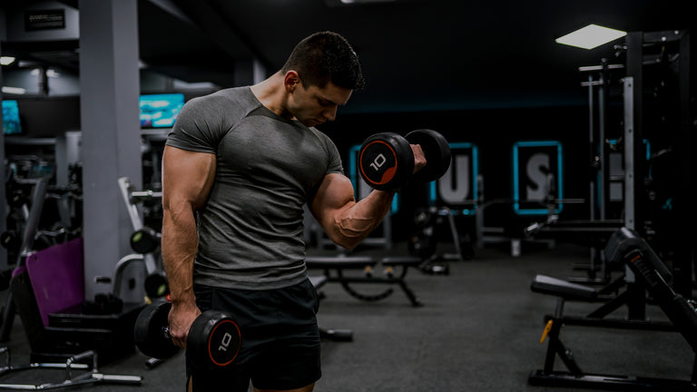 A muscular man in a gym setting, wearing a dark gray t-shirt and black shorts while lifting dumbbells.