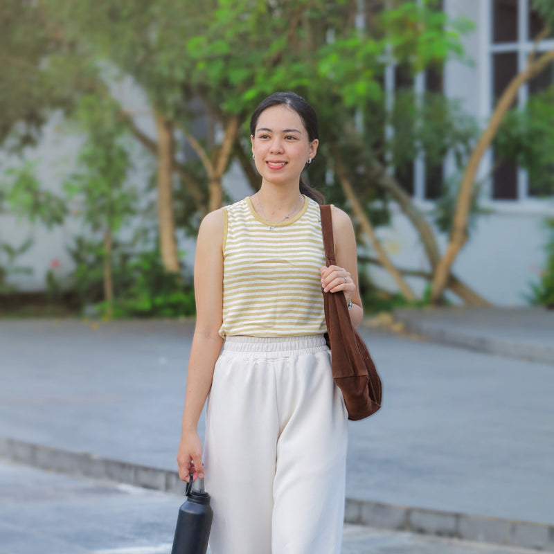 A woman standing outdoors wearing a yellow and white striped sleeveless top, white wide-leg pants, and carrying a brown shoulder bag.