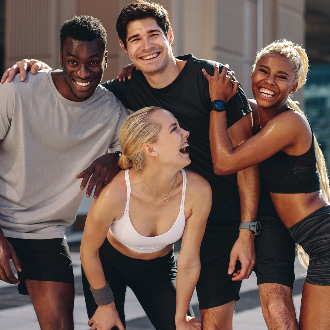 Four smiling people posing together outdoors in athletic wear. Two men and two women are visible from the chest down, wearing black shorts, black and white activewear, and athletic accessories.