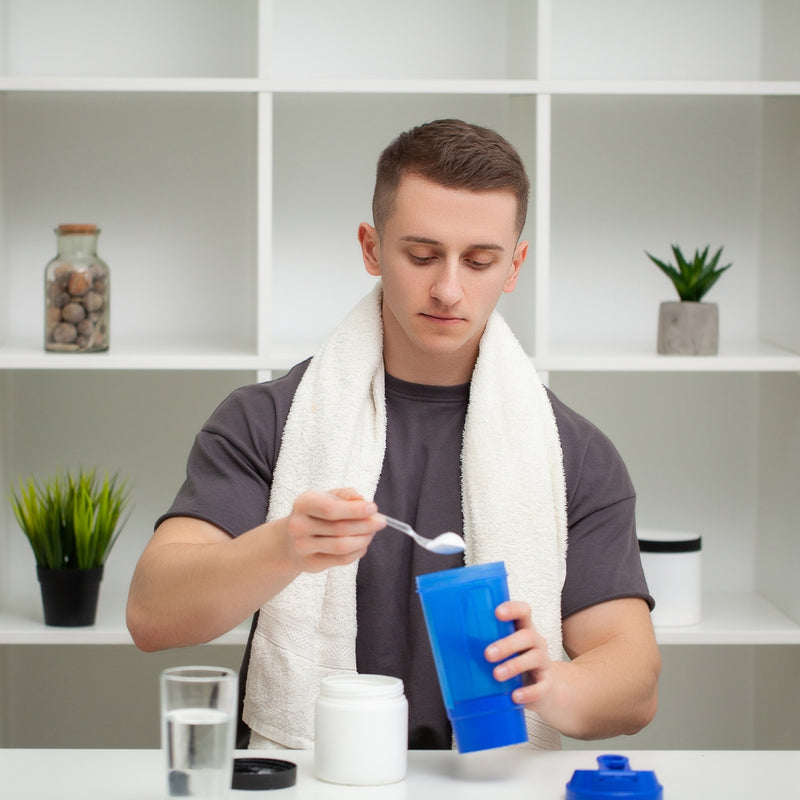 A man wearing a white towel over his shoulders is mixing a blue shaker bottle with a spoon in a kitchen setting.
