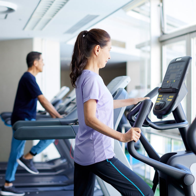 A woman in a light purple t-shirt and black athletic pants uses an elliptical trainer in a bright gym, with a man working out on treadmills in the background.