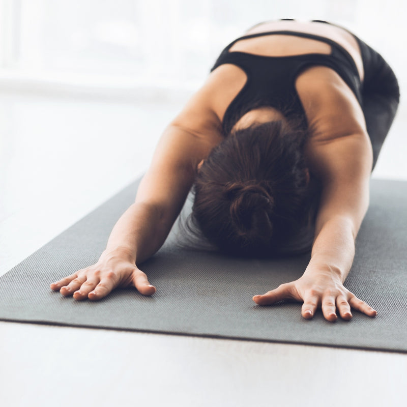 A woman in a black sports bra and leggings poses in a deep forward bend on a dark gray yoga mat in a bright, minimalist room.